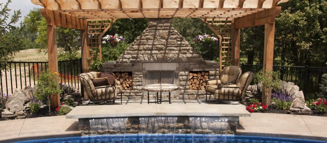 patio pergola with stone fireplace and seating around and a pool with water spilling into it